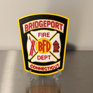 May include: A shield-shaped sign with a black background and yellow trim. The sign features the words "BRIDGEPORT FIRE DEPT CONNECTICUT" in red and white, along with a fire department emblem.