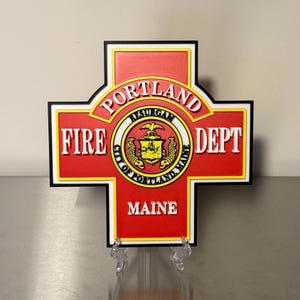 May include: A red and black sign for the Portland Fire Dept, Maine. The sign is in the shape of a cross with the words "FIRE DEPT" on either side of a circular emblem. The emblem has the words "PORTLAND RESURGAM CITY OF PORTLAND MAINE".