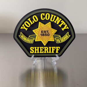 Peut inclure: Un badge noir et jaune du sh&eacute;rif du comt&eacute; de Yolo avec les mots "YOLO COUNTY SHERIFF" et "HONORED TO SERVE" en lettres jaunes. Le badge comporte &eacute;galement une &eacute;toile dor&eacute;e et le texte "EST. 1850".