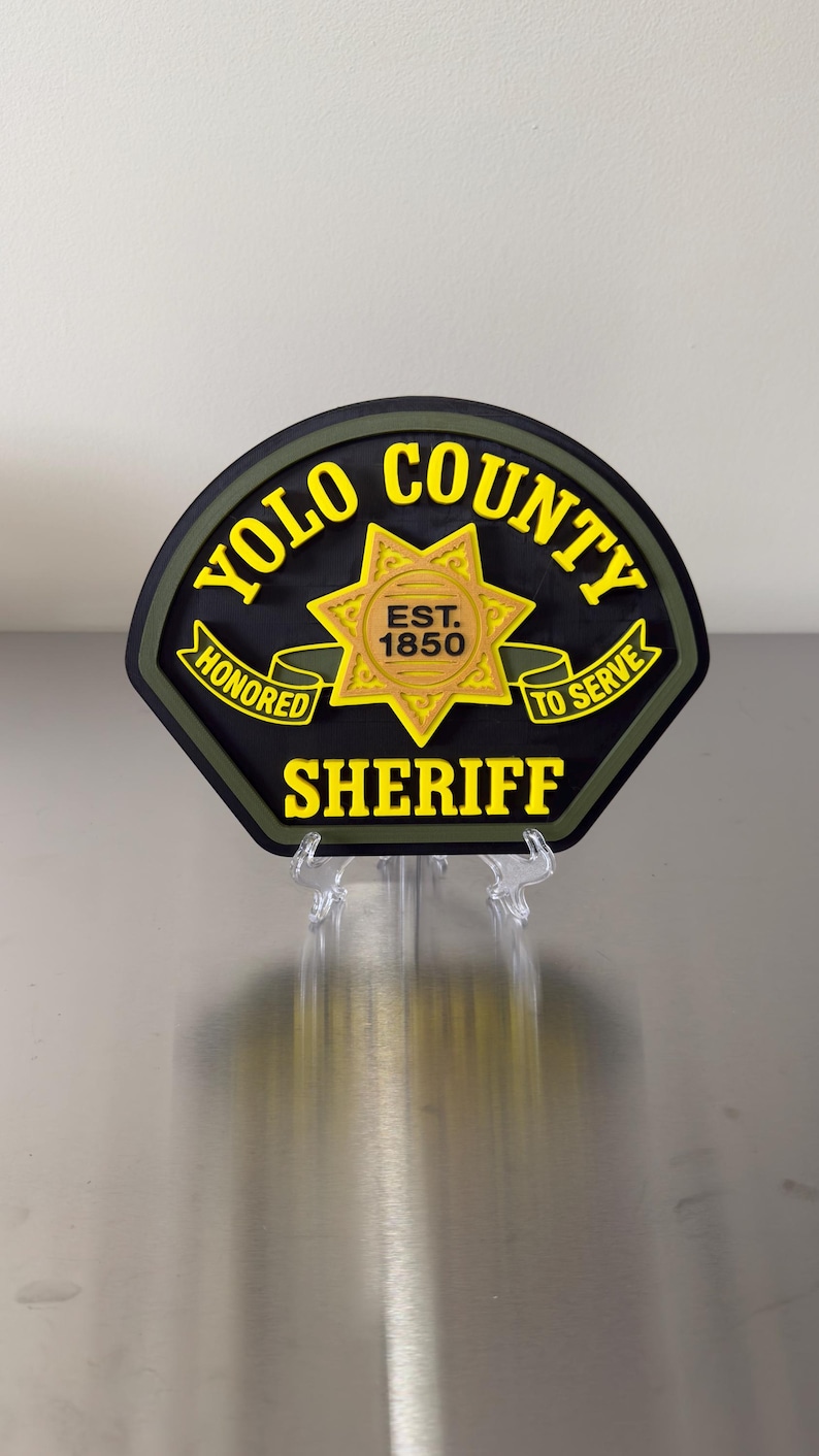 Peut inclure: Un badge noir et jaune du sh&eacute;rif du comt&eacute; de Yolo avec les mots "YOLO COUNTY SHERIFF" et "HONORED TO SERVE" en lettres jaunes. Le badge comporte &eacute;galement une &eacute;toile dor&eacute;e et le texte "EST. 1850".