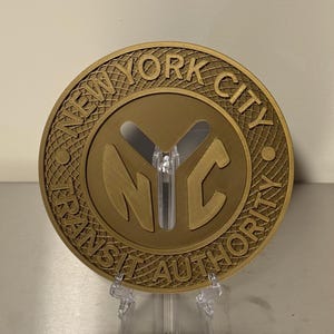 May include: A circular gold-colored medallion featuring the embossed text NEW YORK CITY TRANSIT AUTHORITY around the perimeter and the letters NYC in the center. The background of the medallion has a textured grid pattern.