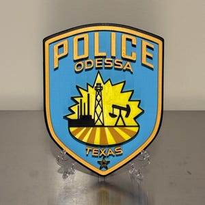 May include: A shield-shaped Odessa Police Texas emblem featuring a light blue background with yellow text and a central graphic of an oil derrick and pump jack over a field. The design includes a black border and a small star at the bottom point.