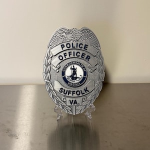 May include: A silver-colored police officer badge with the words "POLICE OFFICER" and "SUFFOLK VA." The badge features a central emblem with the words "COMMONWEALTH OF VIRGINIA". The badge is displayed on a clear stand.