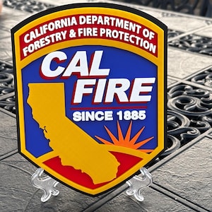 May include: A shield-shaped Cal Fire sign featuring a blue background with a yellow silhouette of California, a red and orange sunburst, and white text reading California Department of Forestry & Fire Protection, Cal Fire, and Since 1885.
