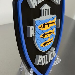 May include: A shield-shaped emblem with the words "WESTERLY POLICE" in gray, 3D lettering. The shield has a black background with a blue border. The center features a blue crest with three yellow fish.