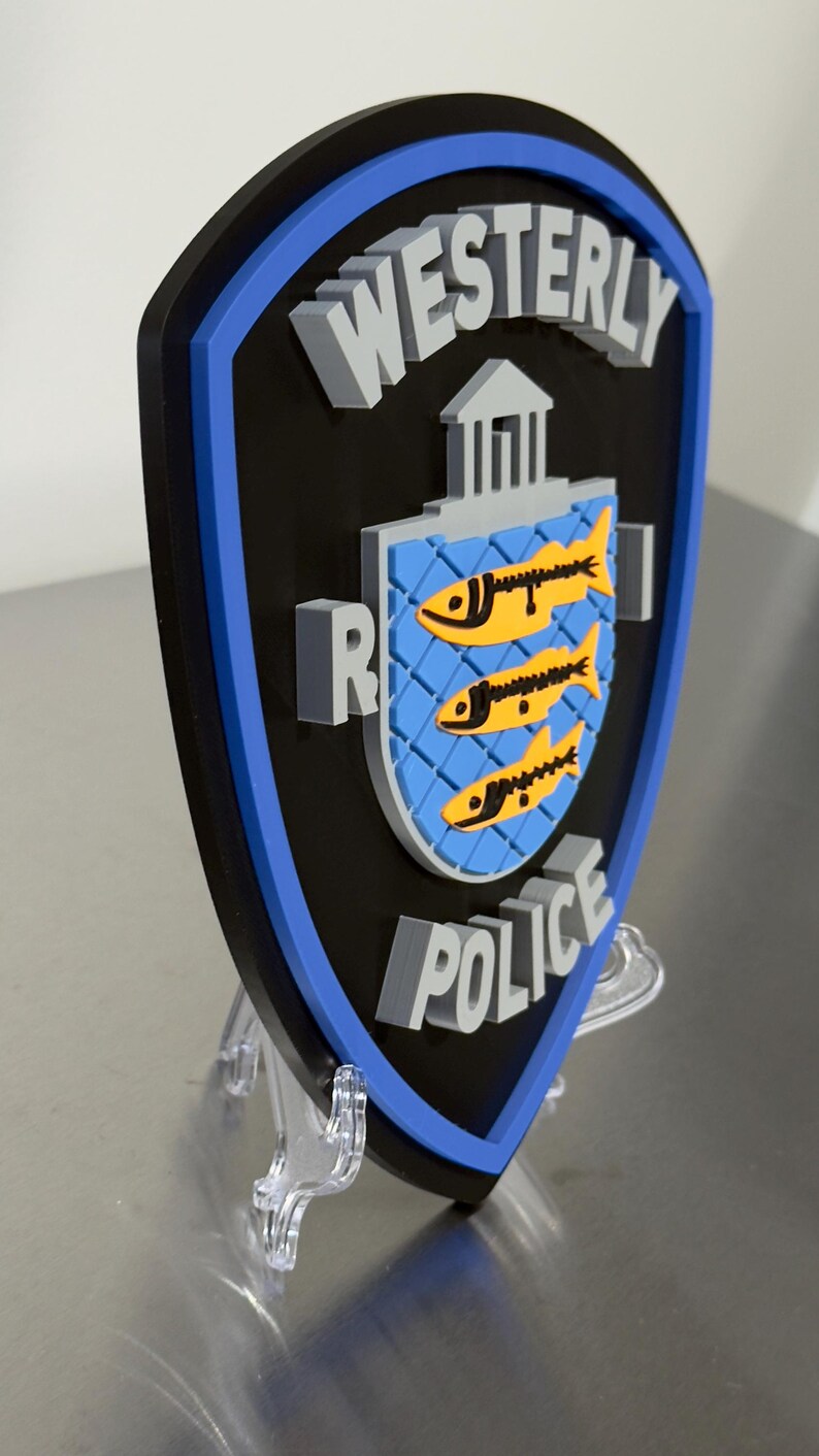 May include: A shield-shaped emblem with the words "WESTERLY POLICE" in gray, 3D lettering. The shield has a black background with a blue border. The center features a blue crest with three yellow fish.