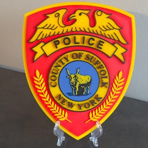 May include: A red and yellow police badge with the words "Police, County of Suffolk, New York" and a yellow eagle with outstretched wings. The badge also features a blue circle with a black bull inside.