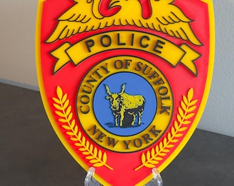 May include: A red and yellow police badge with the words "Police, County of Suffolk, New York" and a yellow eagle with outstretched wings. The badge also features a blue circle with a black bull inside.