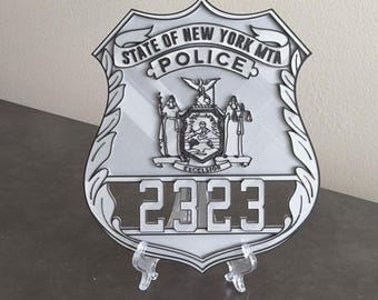 May include: A silver-toned State of New York MTA Police shield badge featuring the text STATE OF NEW YORK MTA, POLICE, and EXCELSIOR. The center displays the New York state seal with two figures and an eagle, with the number 2323 at the bottom.