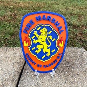 May include: A shield-shaped sign with the words "FIRE MARSHAL" and "COUNTY OF NASSAU N.Y." in blue lettering on an orange background. The center features a yellow lion emblem. Two red flames are on either side of the emblem.