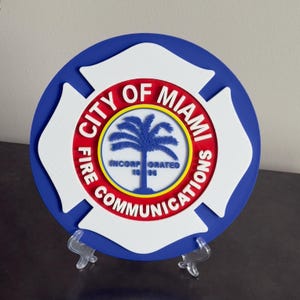 May include: A round, blue, white, and red City of Miami Fire Communications emblem. The emblem features a palm tree in the center, with the words "City of Miami Fire Communications" and "Incorporated 1896" around the edges.