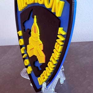 May include: A blue, black, and yellow Madison Police Department emblem. The emblem features the city name in yellow, the words "Police Department" in yellow, and a yellow building outline. The emblem is displayed on a clear stand.