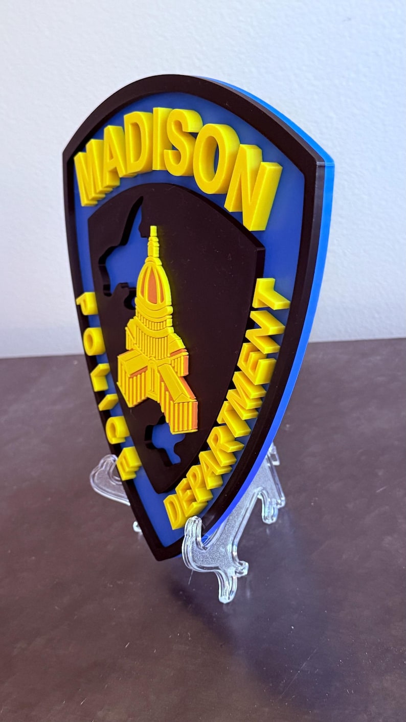 May include: A blue, black, and yellow Madison Police Department emblem. The emblem features the city name in yellow, the words "Police Department" in yellow, and a yellow building outline. The emblem is displayed on a clear stand.