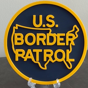 May include: A round, yellow and blue sign with the text "U.S. BORDER PATROL" in yellow letters. The sign features an outline of the United States in yellow.