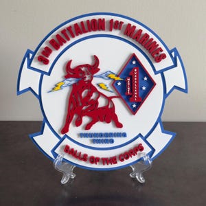 May include: A layered, circular emblem with a red bull, lightning bolts, and text. The top reads "3rd Battalion 1st Marines" in red. A blue and white border surrounds the design. The text "Balls of the Corps" is at the bottom.