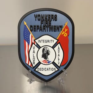 May include: A shield-shaped emblem for the Yonkers Fire Department, featuring the American flag and a red flag with "YFD." The emblem includes the words "Integrity," "Valor," "Dedication," and "Loyalty." The shield is blue with a black border.