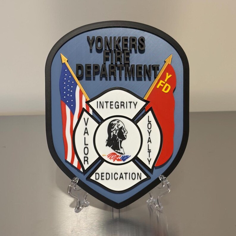 Op de afbeelding: Een schildvormig embleem voor de brandweer van Yonkers, met de Amerikaanse vlag en een rode vlag met "YFD". Het embleem bevat de woorden "Integriteit", "Moed", "Toewijding" en "Loyaliteit". Het schild is blauw met een zwarte rand.