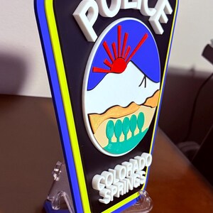 K&ouml;nnte beinhalten: Ein 3D-gedrucktes Polizeibadge mit blauem Hintergrund, wei&szlig;en Buchstaben und gelbem Rand. Das Abzeichen zeigt eine stilisierte Bergszene mit einer roten Sonne und dem Text "POLICE" und "COLORADO SPRINGS".