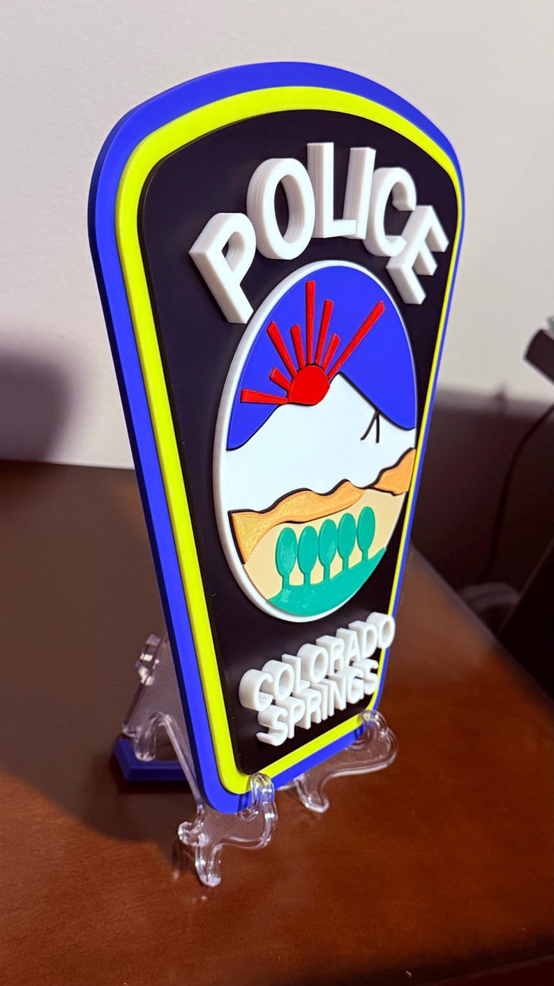 K&ouml;nnte beinhalten: Ein 3D-gedrucktes Polizeibadge mit blauem Hintergrund, wei&szlig;en Buchstaben und gelbem Rand. Das Abzeichen zeigt eine stilisierte Bergszene mit einer roten Sonne und dem Text "POLICE" und "COLORADO SPRINGS".