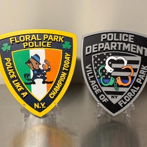 May include: Two shield-shaped plaques. The left plaque is yellow and blue with the text 'FLORAL PARK POLICE' and 'POLICE LIKE A CHAMPION TODAY N.Y.' featuring a leprechaun graphic. The right plaque is grey and black with 'POLICE DEPARTMENT VILLAGE OF FLORAL PARK NY' and a handcuff graphic.