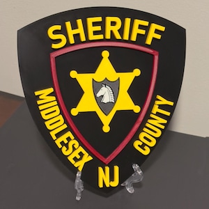 May include: A black shield-shaped plaque with the words "SHERIFF MIDDLESEX COUNTY NJ" in yellow lettering. The center features a yellow star with a gray emblem of a horse's head.