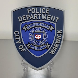 May include: A navy blue police badge with a silver border. The badge reads "POLICE DEPARTMENT" and "CITY OF WARWICK" with the text "TO SERVE AND PROTECT" and "WITH INTEGRITY". The center features a seal with the text "SEAL OF THE CITY OF WARWICK 1642-1963".