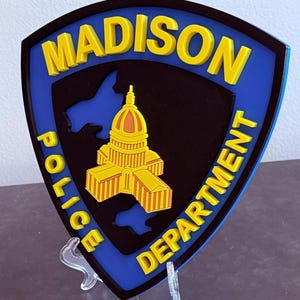 May include: A shield-shaped emblem for the Madison Police Department. The emblem features the city name in yellow, a yellow and orange capitol building, and the words "Police Department" in yellow. The background is black and blue.