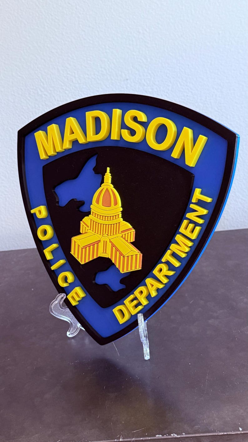 May include: A shield-shaped emblem for the Madison Police Department. The emblem features the city name in yellow, a yellow and orange capitol building, and the words "Police Department" in yellow. The background is black and blue.