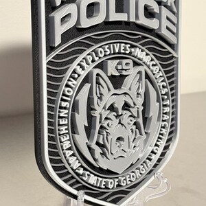 May include: A 3D-printed Woodstock Police badge in black and silver. The badge features the words "Woodstock Police" at the top, with "K-9" and a dog graphic in the center. The badge also includes the text "Explosives Narcotics Tracking" and "State of Georgia".