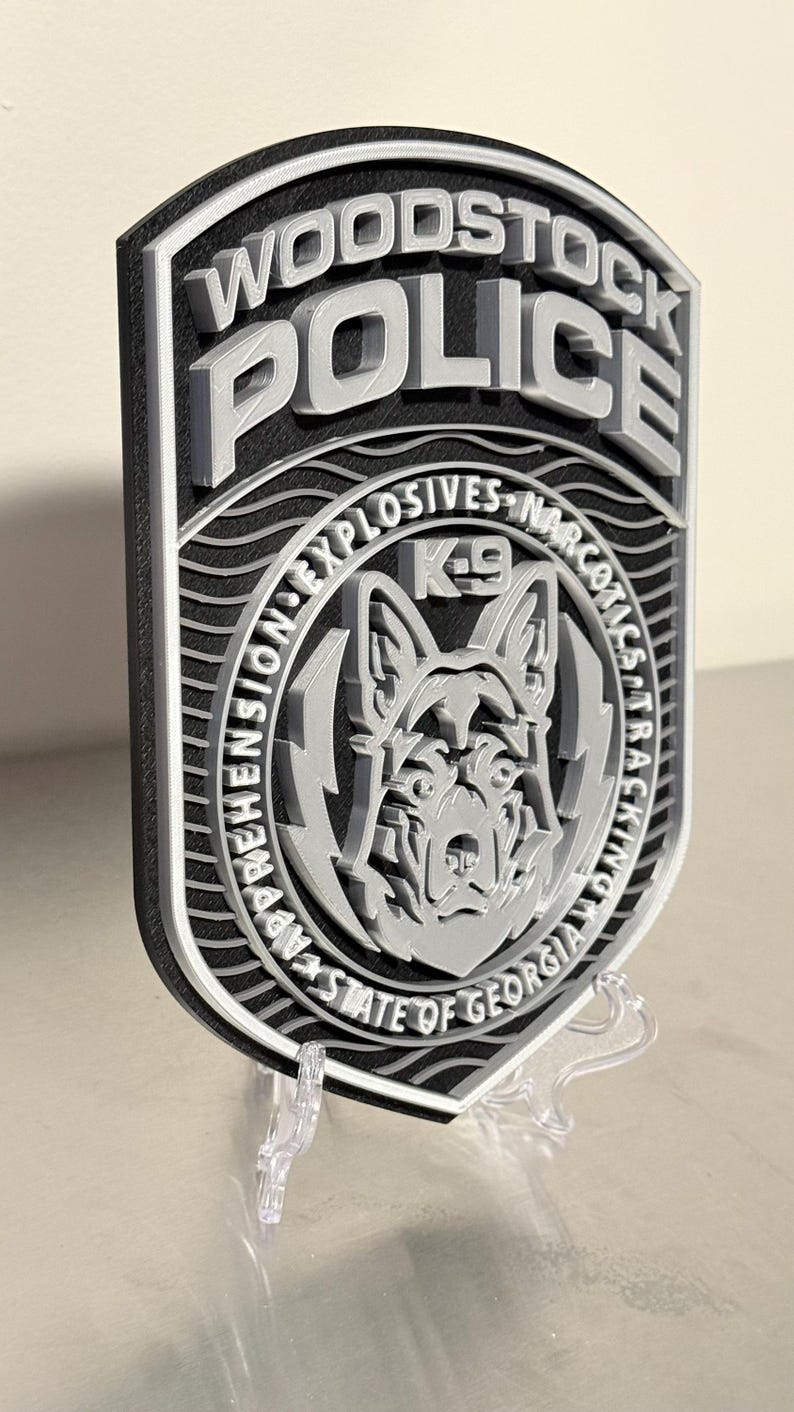 May include: A 3D-printed Woodstock Police badge in black and silver. The badge features the words "Woodstock Police" at the top, with "K-9" and a dog graphic in the center. The badge also includes the text "Explosives Narcotics Tracking" and "State of Georgia".
