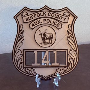 May include: Wooden shield-shaped plaque with the text "SUFFOLK COUNTY AUX. POLICE" and a circular emblem of a bull. The number "141" is displayed below the emblem. The plaque is supported by a clear plastic stand.
