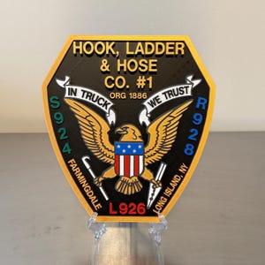 May include: A black and gold emblem with the text "HOOK, LADDER & HOSE CO. #1" and "IN TRUCK WE TRUST". The emblem features an eagle with a shield and the text "FARMINGDALE L926 LONG ISLAND, NY".