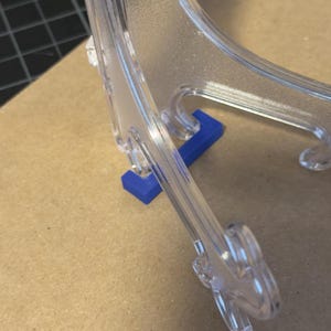 May include: Clear plastic display stand with a blue support piece. The stand is designed to hold items upright. The stand is on a brown surface.