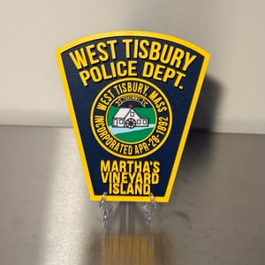 May include: A navy blue and yellow police badge featuring the text "WEST TISBURY POLICE DEPT." and "MARTHA'S VINEYARD ISLAND." The badge has a circular emblem with a building and the text "WEST TISBURY, MASS".