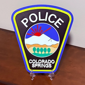 K&ouml;nnte beinhalten: Ein schwarzes und blaues Polizeibadge mit den Worten "POLICE" und "COLORADO SPRINGS" in wei&szlig;en Buchstaben. Das Abzeichen zeigt eine stilisierte Bergkette mit einer aufgehenden roten Sonne dahinter.