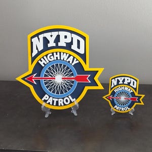 May include: Two NYPD Highway Patrol badges, one larger than the other, are displayed on clear plastic stands. The badges are blue, yellow, and red with a white wheel and arrow.