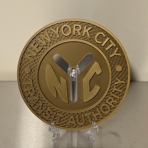 May include: A gold-colored, circular emblem with the text "NEW YORK CITY TRANSIT AUTHORITY" around the edge. The letters "NYC" are prominently displayed in the center. The emblem is mounted on a clear plastic stand.