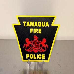 May include: A black and yellow sign with the words "Tamaqua Fire Police" in yellow lettering. The sign has a red emblem in the center. The sign is supported by two clear plastic stands.