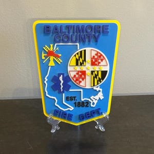 May include: A blue and yellow Baltimore County Fire Dept. sign. The sign features the words "Baltimore County" in blue, and "Fire Dept." in blue. The sign also has a Maryland flag design and a star of life symbol.