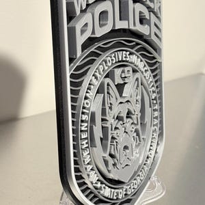 May include: A layered, 3D-printed police badge for the Woodstock Police Department. The badge features the words "Woodstock Police" and "K9" with a dog graphic. The badge is gray and black and is displayed on a clear stand.