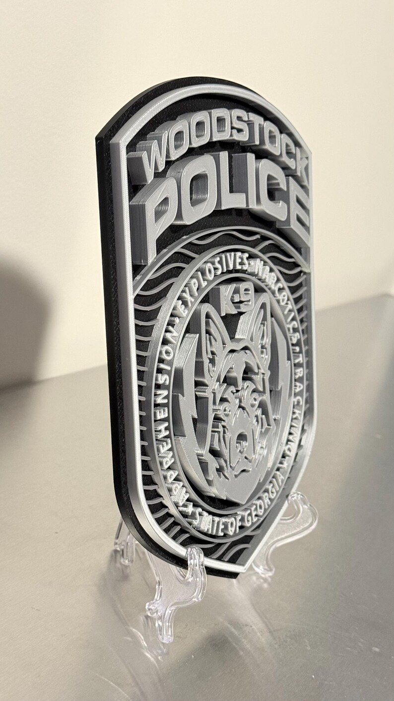 May include: A layered, 3D-printed police badge for the Woodstock Police Department. The badge features the words "Woodstock Police" and "K9" with a dog graphic. The badge is gray and black and is displayed on a clear stand.