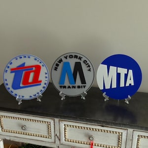 May include: Three round, decorative plaques with logos for the New York City Transit Authority, the MTA, and the New York City Transit. The plaques are white, gray, and blue with red, black, and white lettering.
