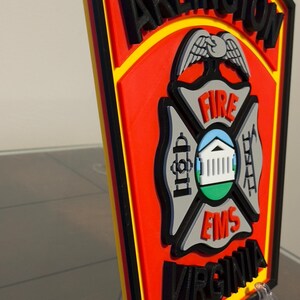May include: A 3D printed, red, yellow, and black Arlington, Virginia Fire and EMS patch with a black eagle and the words "FIRE" and "EMS" in white. The patch is on a clear plastic stand.