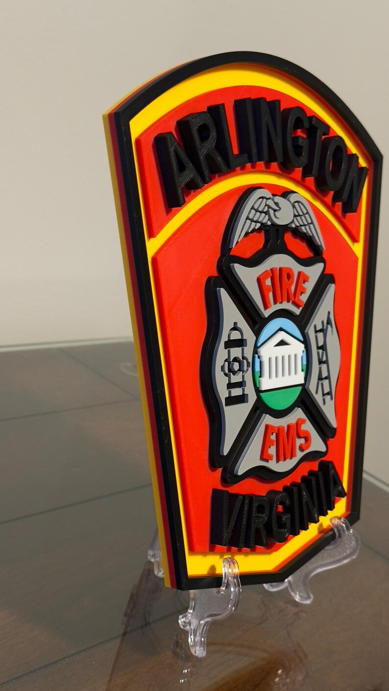 May include: A 3D printed, red, yellow, and black Arlington, Virginia Fire and EMS patch with a black eagle and the words "FIRE" and "EMS" in white. The patch is on a clear plastic stand.