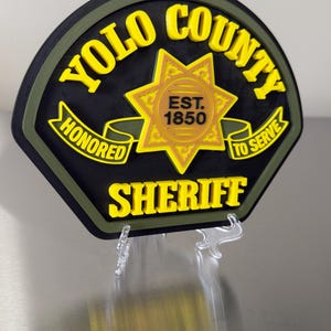 Peut inclure: Un badge de sh&eacute;rif du comt&eacute; de Yolo noir et jaune avec une &eacute;toile dor&eacute;e au centre. Le badge porte les mots "YOLO COUNTY SHERIFF" et "HONORED TO SERVE" en lettres jaunes. Le badge est mont&eacute; sur un support transparent.