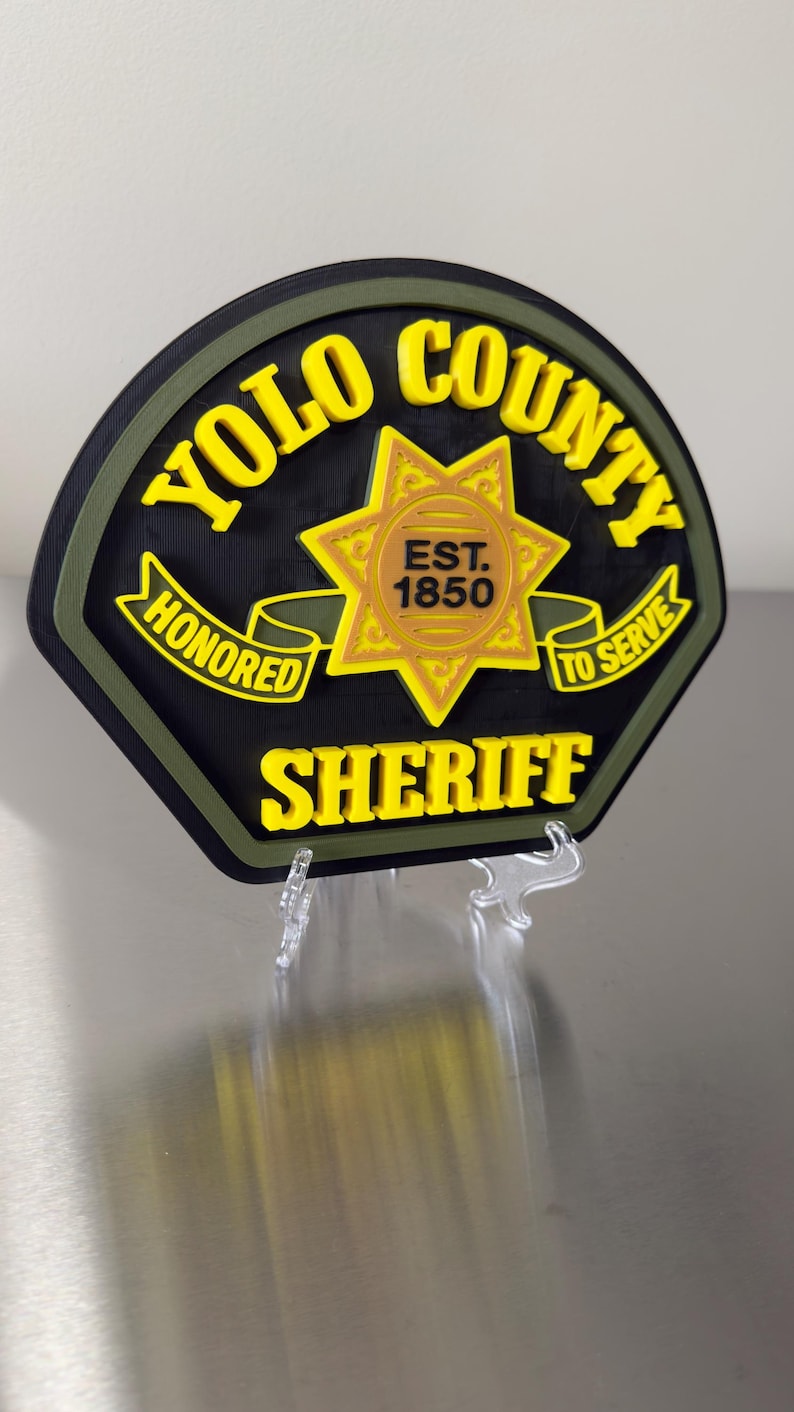 Peut inclure: Un badge de sh&eacute;rif du comt&eacute; de Yolo noir et jaune avec une &eacute;toile dor&eacute;e au centre. Le badge porte les mots "YOLO COUNTY SHERIFF" et "HONORED TO SERVE" en lettres jaunes. Le badge est mont&eacute; sur un support transparent.