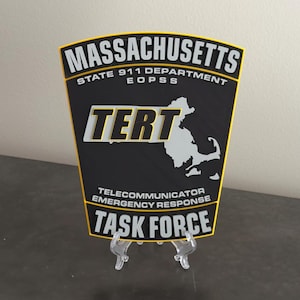 May include: A black patch with yellow trim, featuring the text "MASSACHUSETTS STATE 911 DEPARTMENT EOPSS TERT TELECOMMUNICATOR EMERGENCY RESPONSE TASK FORCE" and a white outline of the state of Massachusetts.