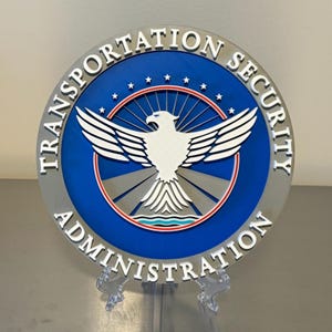 May include: A round Transportation Security Administration (TSA) sign. The sign features a blue center with a white eagle emblem and the text "TRANSPORTATION SECURITY ADMINISTRATION" in white. The outer ring is gray.