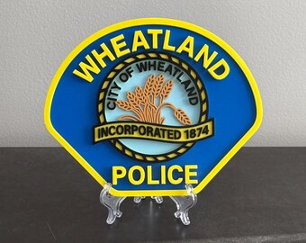 May include: A blue and yellow shield-shaped sign featuring the text WHEATLAND at the top and POLICE at the bottom. The center displays a circular emblem with wheat stalks and the text CITY OF WHEATLAND INCORPORATED 1874 on a banner.