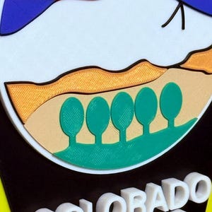 K&ouml;nnte beinhalten: Ein 3D-gedrucktes Schild mit blauem Hintergrund und einer wei&szlig;en Bergszene mit einer aufgehenden roten Sonne. Das Schild tr&auml;gt die Aufschrift "COLORADO SPRINGS" in wei&szlig;en Buchstaben.